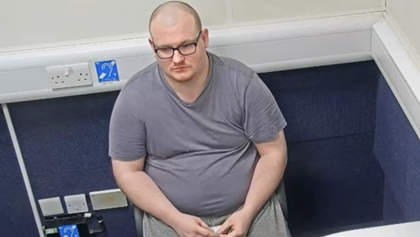 Matthew Webb is sitting in a police interview room. He is bald, is wearing dark glasses and a grey top.