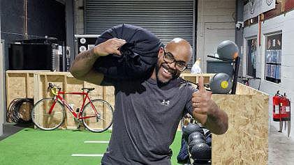 Daniel Jude wearing a grey t-shirt carrying a 50kg sandbag on his shoulder holding his thumb up smiling in a gym