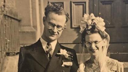 A black and white photo of a man with short, curly hair and glasses, wearing a black suit, white shirt and a tie. He has a flower pinned to his jacket and is smiling at the camera. Next t him is a woman wearing a white lacey dress, a white veil and a head piece and glasses and is also smiling at the camera.