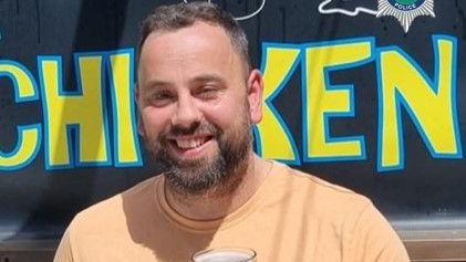 A man with a dark brown beard and short dark hair smiles showing his teeth to the camera. He wears a light orange top and appears to have a beer glass in front of his chest. Behind him are yellow letters with a blue outline spelling "chicken"