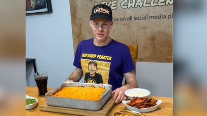 A still from a video, a man sits behind a massive cottage pie in a tray with side plates full of food beside him. He wears a cap and t-shirt which says 'Wayward Feaster'