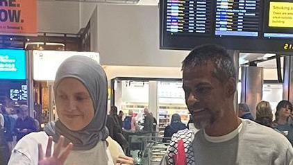 A man and a woman stand side by side, looking to the side of the camera. She wears a hijab and is holding her palm out in the air. He is wearing a grey jumper with a white T-shirt underneath. He has short, black and grey hair.