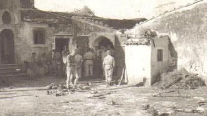 An old photograph from World War Two. There are many soldiers gathered around a wall which leads into an open doorway. The sun is shining casting long shadows and the soldiers are sitting and standing in various points of shade, many of them with their backs turned from the camera.