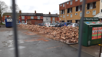 A large pile of rubble, formerly garages in front of a row of cars.  There are flats and houses in the background