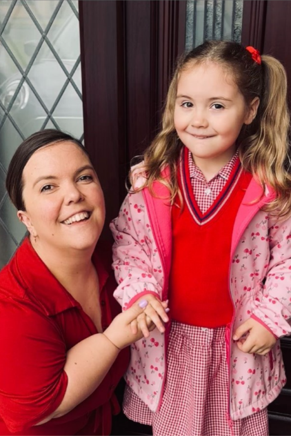 Hanna is kneeling next to Darcy a the inside of a front door with a decorative lead frame window. Darcy has long blonde pig tail hair and is smiling for the cameral with a pink winter jacket and red jumper. Mum has long tied back brown hair and is smiling