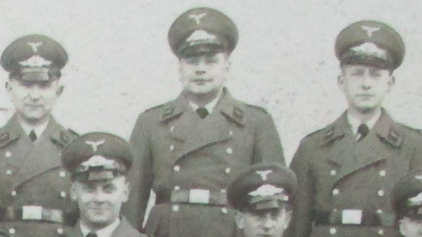 A man poses for a group photograph, wearing a double-breasted uniform jacket and a peaked cap.