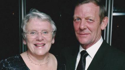 Mary and Bryan Andrews. Both are looking at the camera - Mary has short grey hair and is wearing glasses, a sliver necklace and a black dress. Bryan has brown hair and is wearing a black suit and tie.