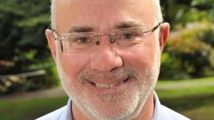 A man with glasses smiling at the camera in a garden