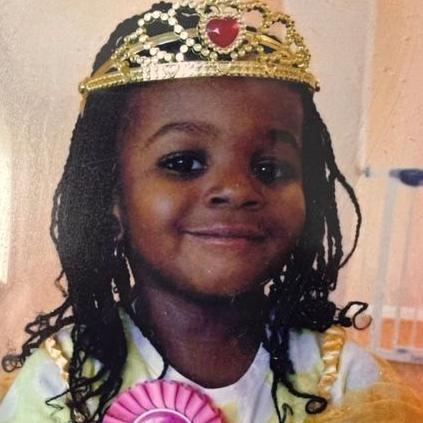 Elianne Andam is smiling. She is a young girl in the photo and is dressed up wearing a gold crown with a pink heart on it. She is wearing a pink rosette.