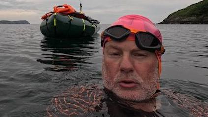 David Higson in a pink swimming cap in the sea around the Isle of Man, with a raft pictured behind him