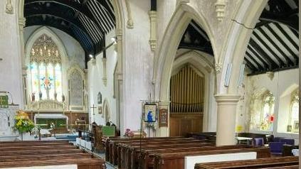 Rows of wooden pews can be seen inside the church, with the altar at the front and a number of stained glass windows. The walls are cream-coloured with a pillar between one of the rows of pews. 