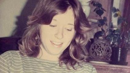 Shirley has brown hair and is wearing a striped sage top. She is smiling and looking downwards