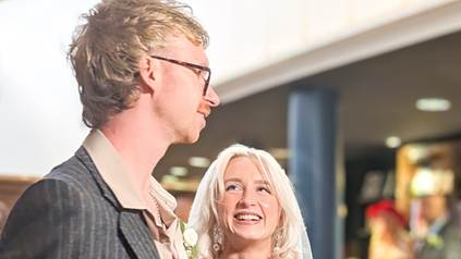 A man with short light brown hair, a salmon-coloured shirt and pinstripe grey suit jacket and smiling while standing next to a woman with long blonde hair and a wedding veil. The main foyer area of Storyhouse can be seen behind them.