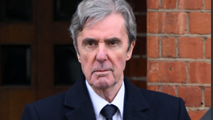 John Stapleton, wearing a dark suit and tie with grey hair, looks straight ahead in a sombre picture