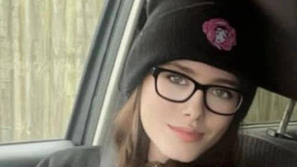 A head and shoulders image of a smiling woman sitting in the front seat of car. She has long brown hair and is wearing black framed spectacles and a black beanie. Her face is turned towards the camera, with the side window behind her.