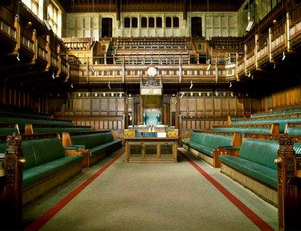 An empty House of Commons. It is a wooden interior with many rows of green benches.