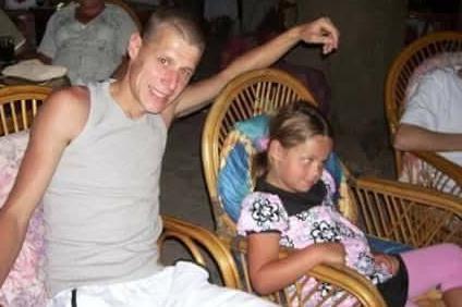 A young man wearing a grey vest and white shorts is sitting in a wicker chair and leaning towards his young daughter, who is wearing a flowery pink dress and sitting in a similar chair. Both are smiling.