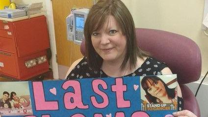 Rebecca Oldham sits in a purple chair in a hospital, with medical equipment visible behind her. She is looking into the camera and holding a blue sign with pink letters reading: "Last chemo treatment".