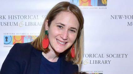 Wesley LePatner poses at New York Historical Society Museum event. She is smiling and wearing a dark blue shirt and blazer and and large dangling red and green earrings. In the background is a backdrop for the museum showing its logo. 