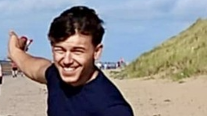 A young man with dark hair and a dark blue t-shirt smiles as he points behind him while standing on a beach.