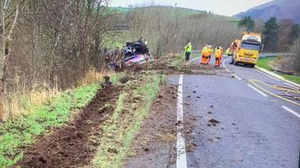 A tanker is on its side on the side of a road surrounded by hills. There are deep track marks on the side of the road leading to the vehicle. A number of people wearing high vis jackets are at the scene