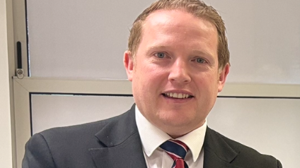 A headshot of a man with short, light brown hair and blue eyes wearing a grey suit with a blue and red striped tie. 