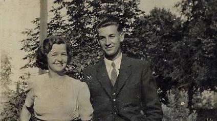 A black and white photo of Grace and Les Gould wearing smart clothing for a photograph. They are both smiling at the camera. There are trees and plants behind them.
