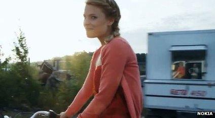 A girl rides her bicycle. A reflection in a window shows that the video was recorded by a professional camera operator riding in a van.