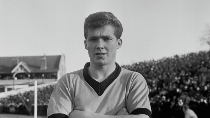 A black and white image of footballer Terry Wharton at a game in 1961. The photo is a close-up of Wharton, who is crossing his arms across his chest, while standing on a football pitch. The crowd is visible behind him.