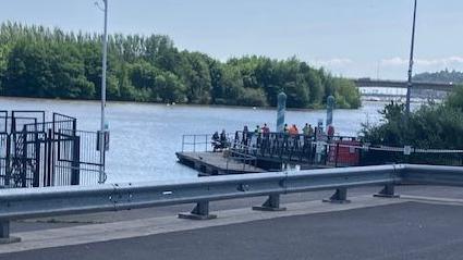 A large body of water with a platform on which several people are stood in a crowd. In the background are trees and in the foreground is a road with a metal barrier.