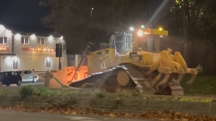 A yellow mechanical digger in a street at night with a white building in the background