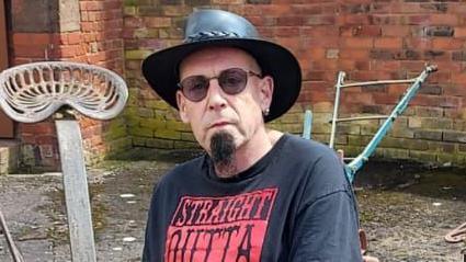 Man sitting outside wearing a black hat, sunglasses and a black top with red writing looks into the camera. 