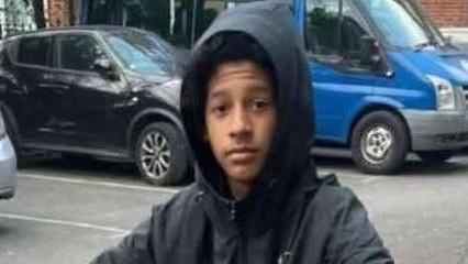 Picture of the teenager looking into the camera, wearing a black hooded jacket. There are car parking spaces behind him with a black car and blue van in two of the spots. 