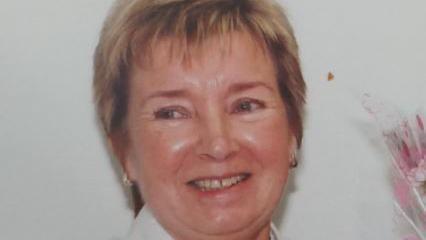 Mary Kinsey is smiling. She has short blonde hair. She looks away from the camera as she stands in front of a white wall with some pink flowers to her right.