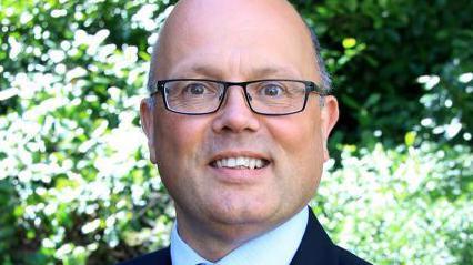 A smiling Andrew Wickham. He is wearing a suit and dark-rimmed glasses.