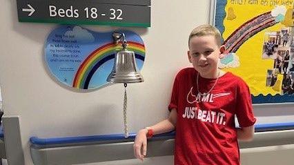 Harry wearing a red t-shirt sayig AML just beat it as he rings the bell in Royal Manchester Children's Hospital to mark the end of his treatment.