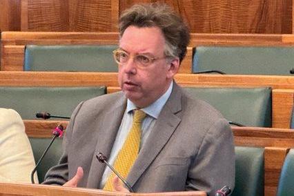 A man sat on a bench in a council meeting, speaking into a microphone. He is wearing a grey suit jacket, a white shirt and a yellow tie, and has yellow round glasses and greying brown short hair.