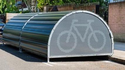 A large, curved metal structure placed on the side of a street The front panel has a bicycle symbol. Behind the unit is a brick wall and residential buildings with greenery.