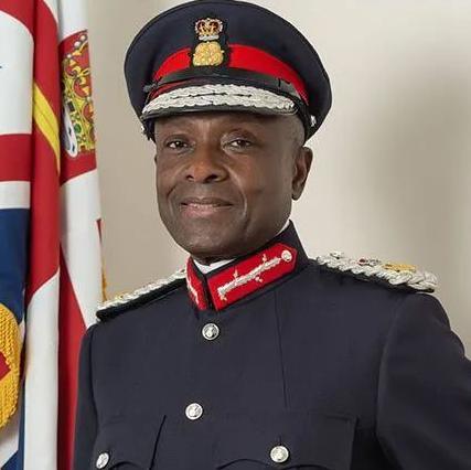 A man in military uniform stood next to a flag. The uniform is navy blue with white shoulder straps and red collar strips. The man's cap is navy blue with a red band and white embroidery on the peak. The uniform has five silver buttons