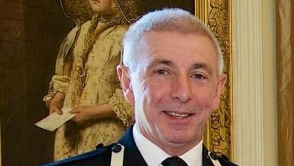 A picture of a man smiling at the camera. He is wearing a white suit shirt, black tie with a black blazer. He has grey hair and is stood in front of a picture. 