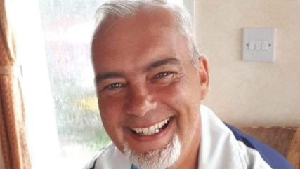 A photograph of Christopher Owens, a middle-aged white man who is balding but has some white hair in a comb-over style and a grey goatee. He has dark brown eyebrows. Mr Owens is smiling and appears to be sitting in an armchair in front of a frosted window.