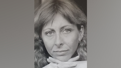 A black and white close up portrait photo of a woman with a fringe and curly light hair. She has her hands interlaced under her chin and is wearing a ring and hoop earrings.