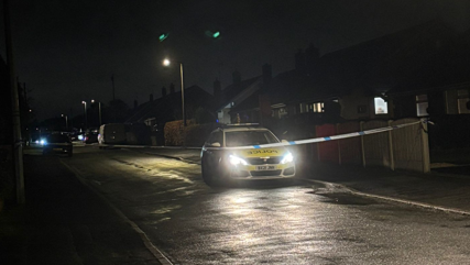 A police car is parked at the side of a residential road, with its headlights on. It is dark, and there is police tape strung across the road.