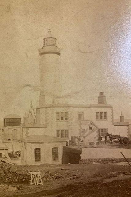 A faded black and white picture of the Corsewall Lighthouse. The tower is in the background with buildings in front of it.