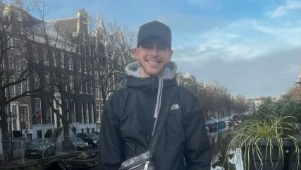 Robert Tyler-Jones looking and smiling at the camera, dressed in a black jacket and wearing a bag and a cap. In the background there's a canal and houses on the side. He's standing on the bridge.
