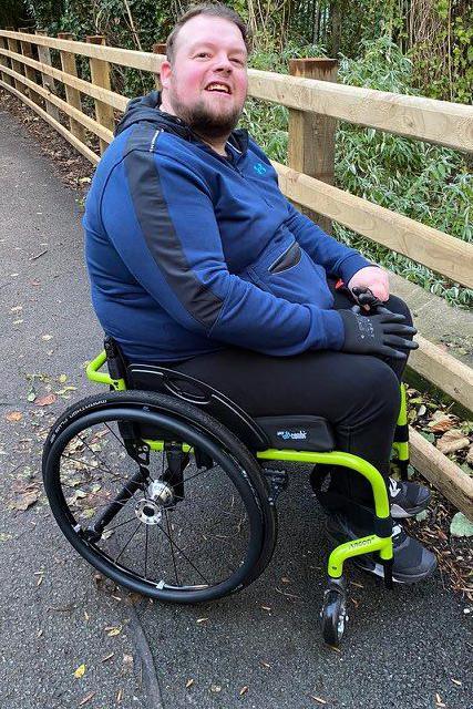 Kieren is sitting in a florescent green wheelchair in a country park. 
He is a white man, sporting a beard and hair swept backwards. 
Kieren is looking at the camera with a smile. 