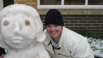 A young woman wearing a black beanie and a big white coat smiles as she stands beside a snow woman in front of a building, potentially a classroom. There is snow on the grass behind her.