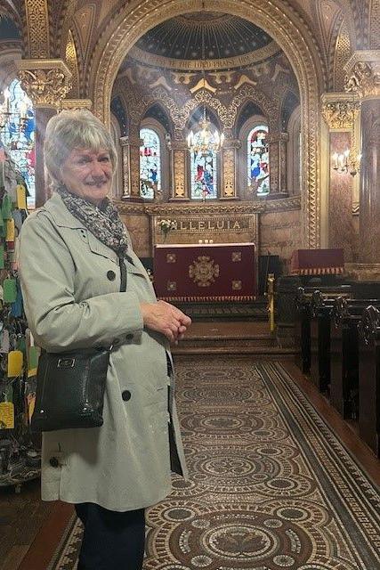 A smiling older woman stands in the aisle of a richly decorated chapel. She was wearing a light green coat.