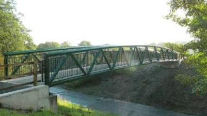 A green-railed footbridge spanning a small waterway, pictured in computerised form. There are green trees on either side and it is light.