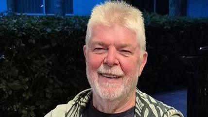 William Cherry is smiling at the camera. He has short, white hair and a white beard. He is wearing a hoodie with a camouflage pattern inside and is standing in front of a hedge and a house.